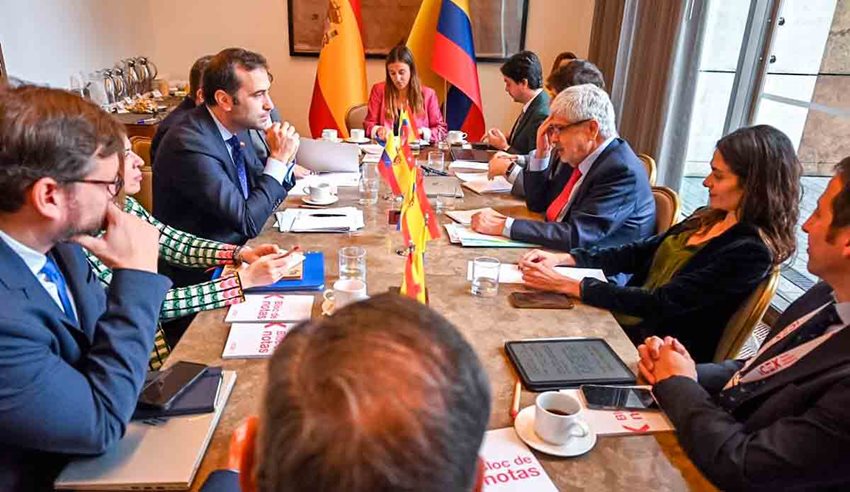 Personas sentadas alrededor de una mesa, con banderas de España y Colombia en el fondo, teniendo una reunión.