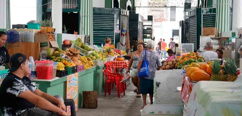 Descripción: Personas caminando en medio de puestos de frutas en una plaza de mercado.